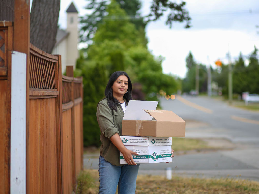 A portrait of a young woman standing in a front yard holding a pile of boxes.