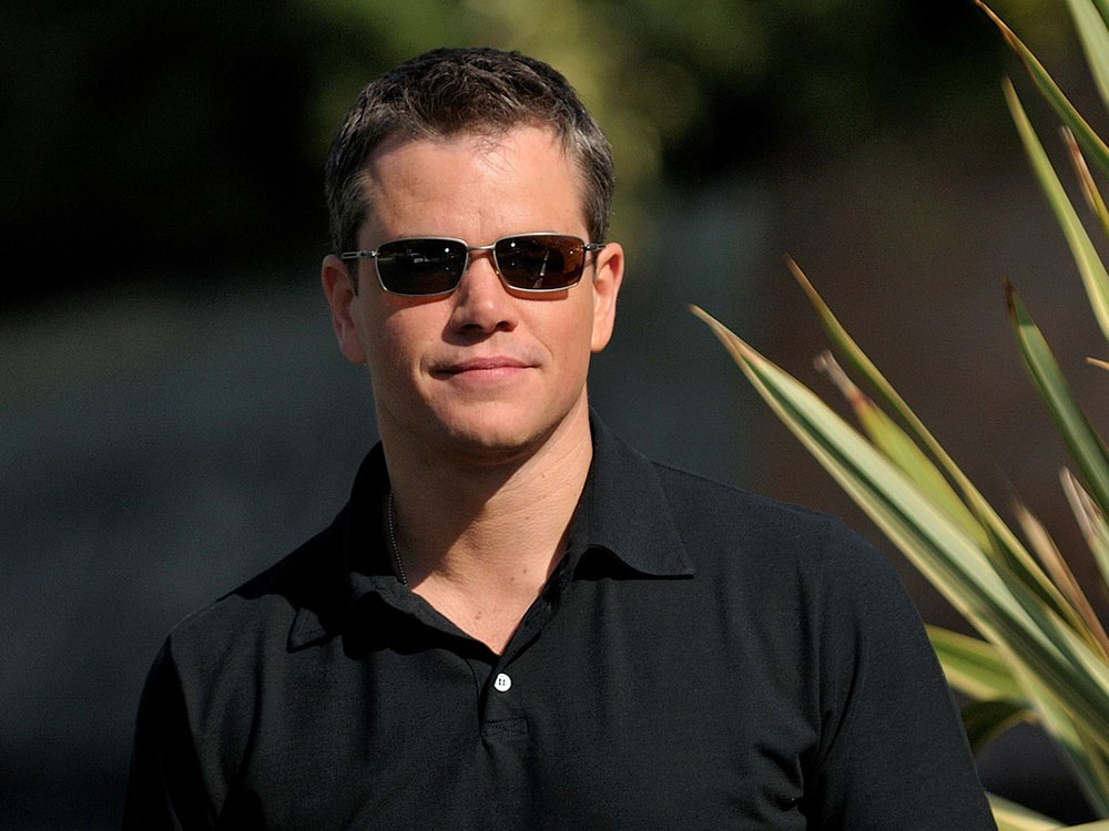 Actor Matt Damon, in sunglasses, stands in front of black background and palm leaves.