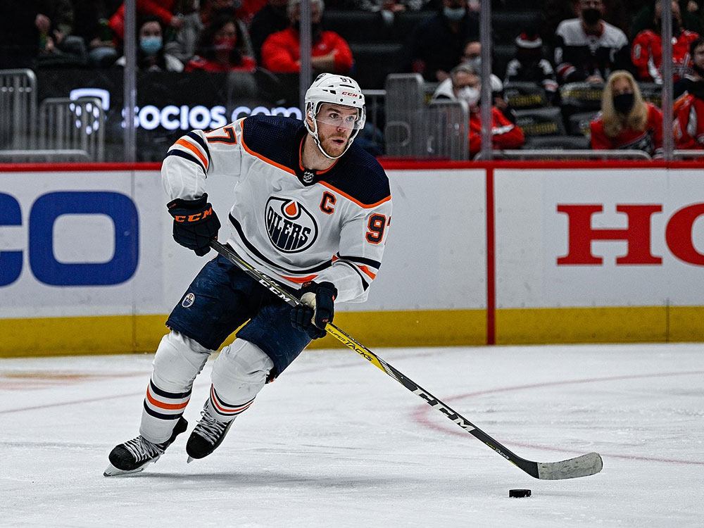 A professional hockey player, Connor McDavid, wearing a white with blue and orange Edmonton Oilers uniform, looks up ice with the puck just off the end of his stick.