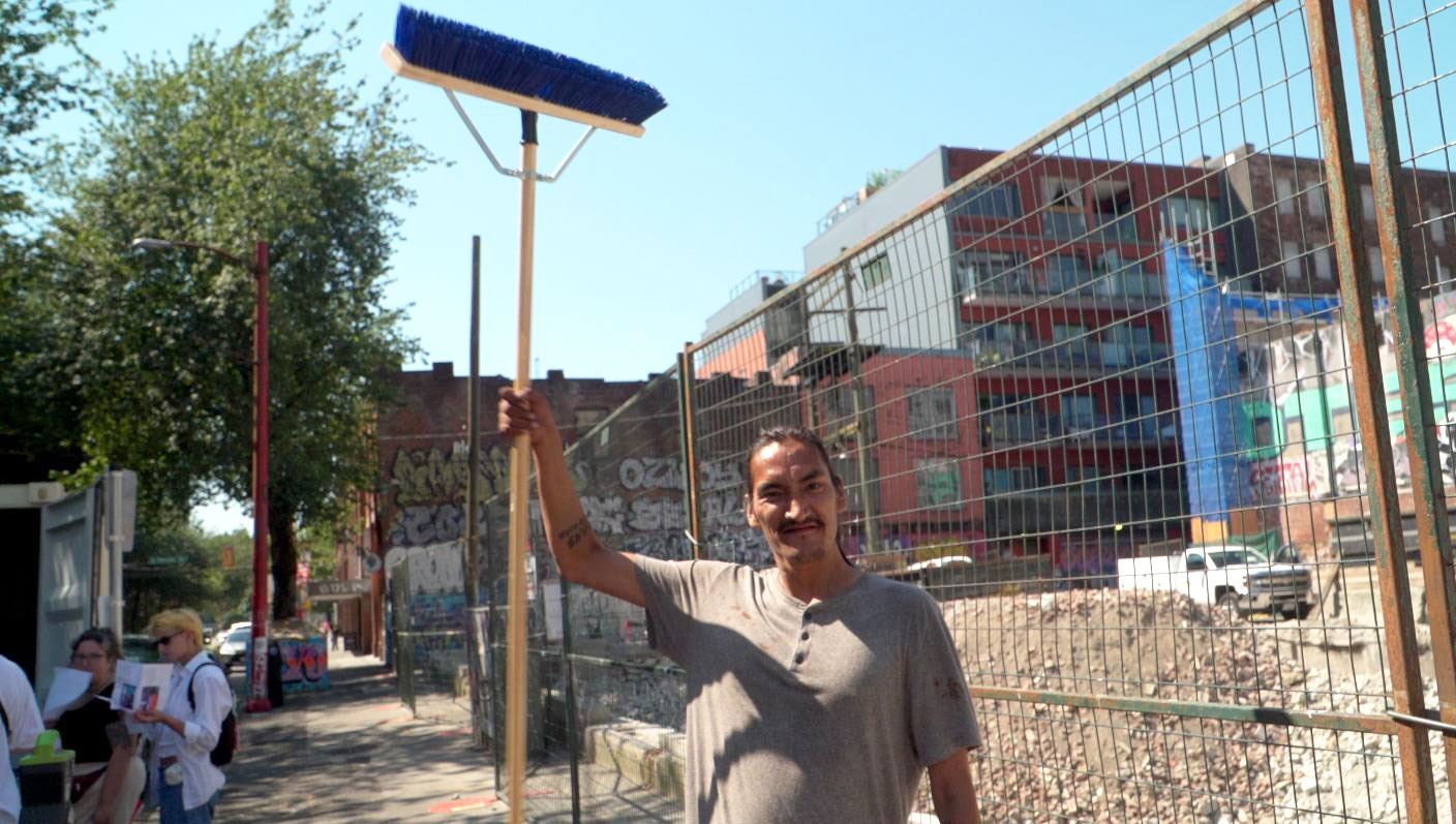 An Indigenous man named Aaron stands on the sidewalk in front of a construction site, holding an industrial-sized janitorial broom over his head.