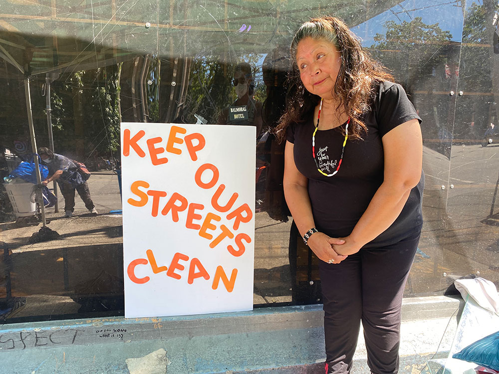 An Indigenous woman, Mama Marcy, is standing to the right of a handmade sign, white posterboard with orange letters, that reads “Keep our streets clean.” Marcy is wearing black and a red, black and white beaded necklace in the colours of the medicine wheel.