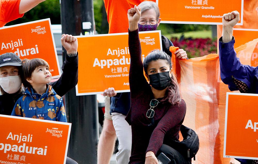 Wearing a black shirt, pants and mask, dark-haired Anjali Appadurai raises a fist surrounded by volunteers holding orange federal NDP election signs bearing her name.