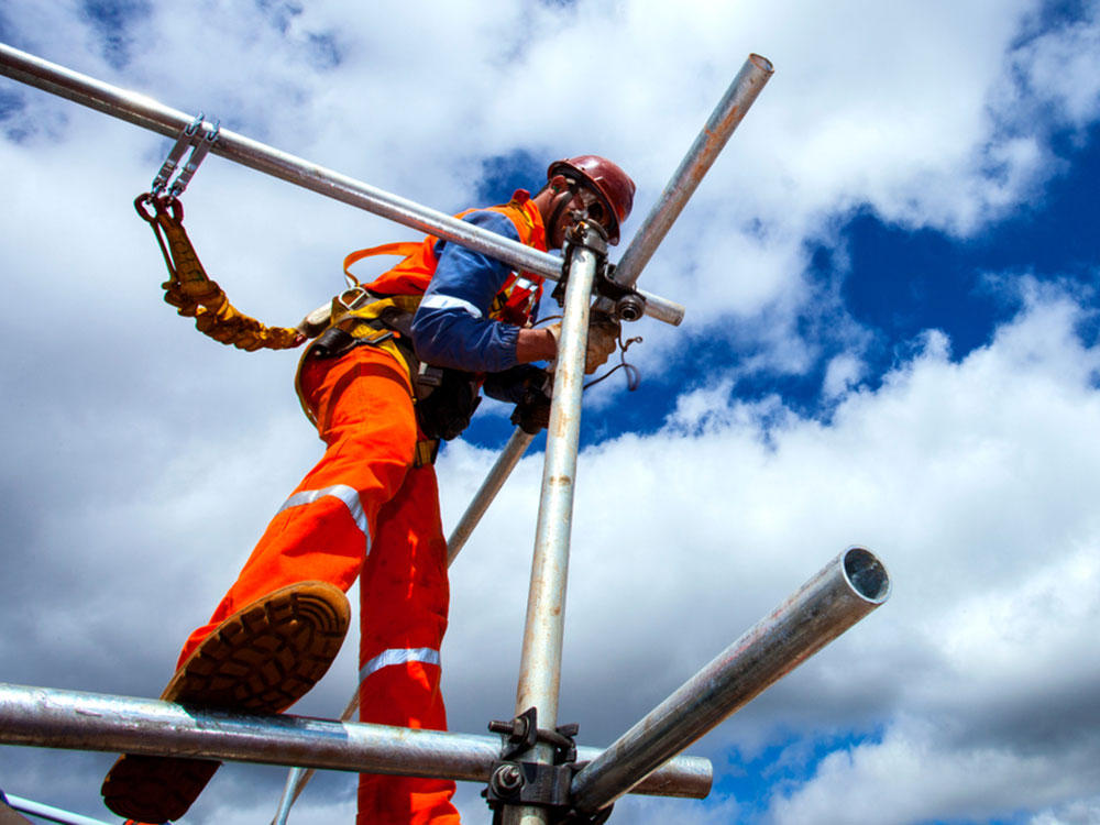 Seen from below, a helmet-wearing construction worker in blue and orange work clothes erects a platform made of metal tubing with a bright blue sky and white clouds behind.