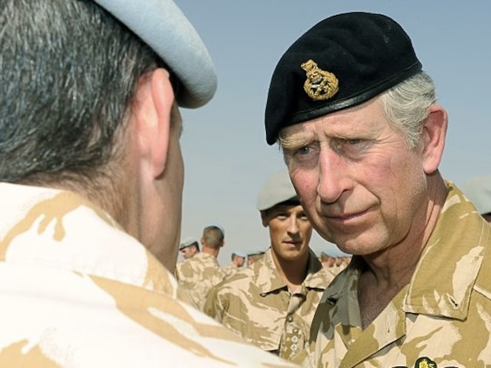 Prince Charles wears a military camouflage uniform and black beret as he speaks with a soldier.