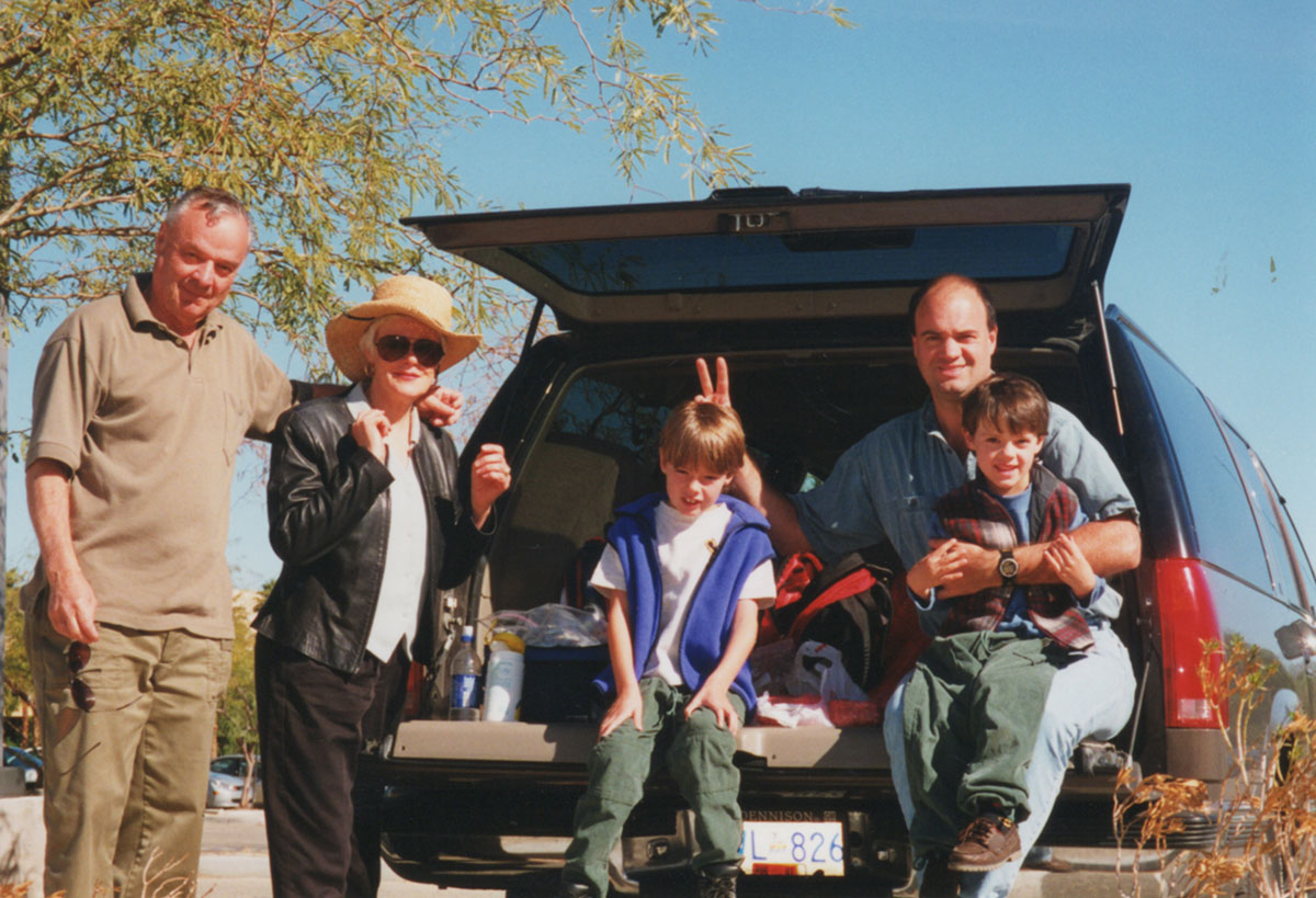 Five people stand in front of the open back of a sports utility vehicle on a sunny day. Glen Fawkes is wearing a blue shirt and holding one of his sons in his lap. He poses jokingly for the camera, holding a peace sign behind his son Kyle’s head. Kyle is wearing a blue vest.