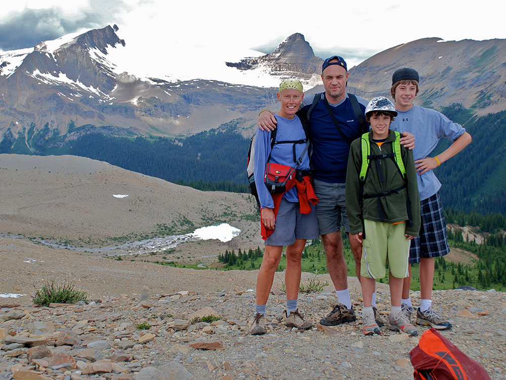 A family of four stands on a hiking trail with a sweeping mountain vista behind them. They are dressed in athletic apparel in blue and green tones. They are standing together to the right of the frame, looking at the camera and smiling. 