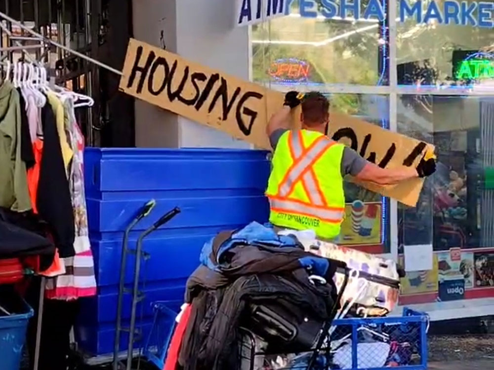 A man in a neon yellow work vest takes down a handmade cardboard sign that reads ‘Housing now’ on East Hastings Street in Vancouver’s Downtown Eastside. The worker is wearing black gloves and his back is turned to the camera. The sign is against the wall of a convenience store. Around him are large blue containers, clothing on racks and piles of bedding.