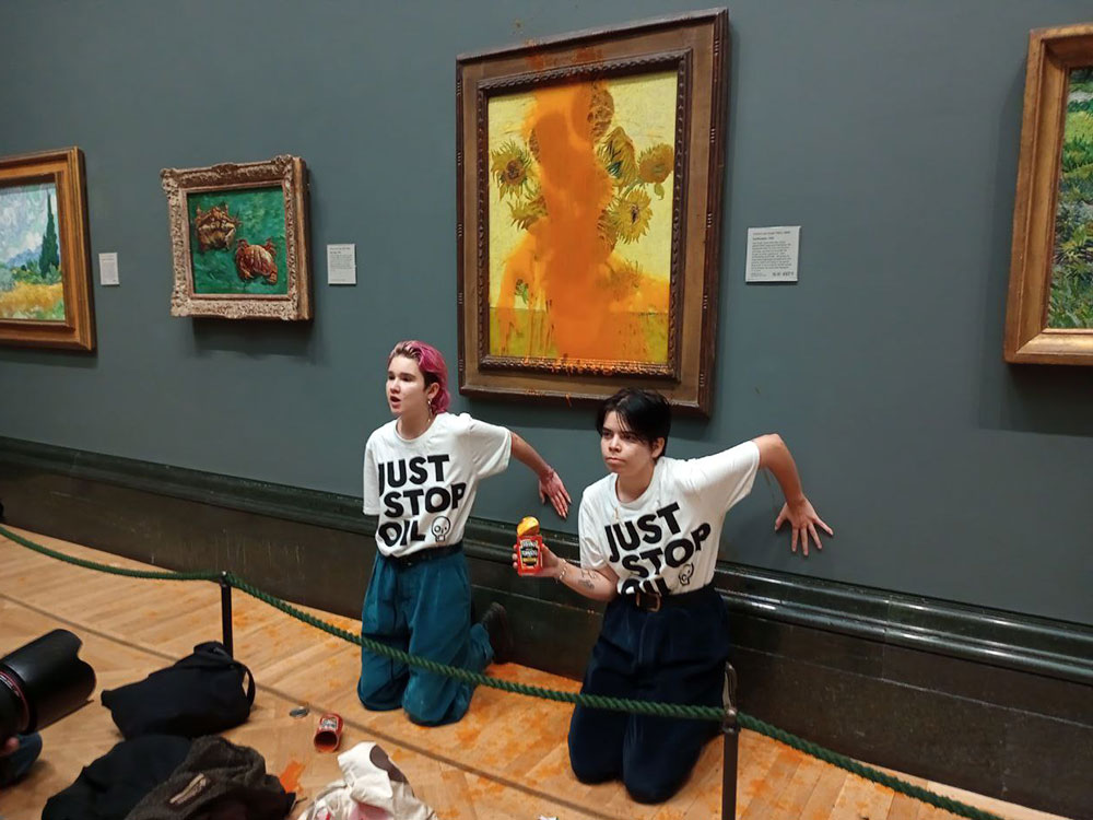 Two people wearing Just Stop Oil T-shirts kneel in front of Vin-cent van Gogh’s painting, “Sunflowers,” which is protected by plexi-glass. The plexiglass is covered in a large splatter of tomato soup. 