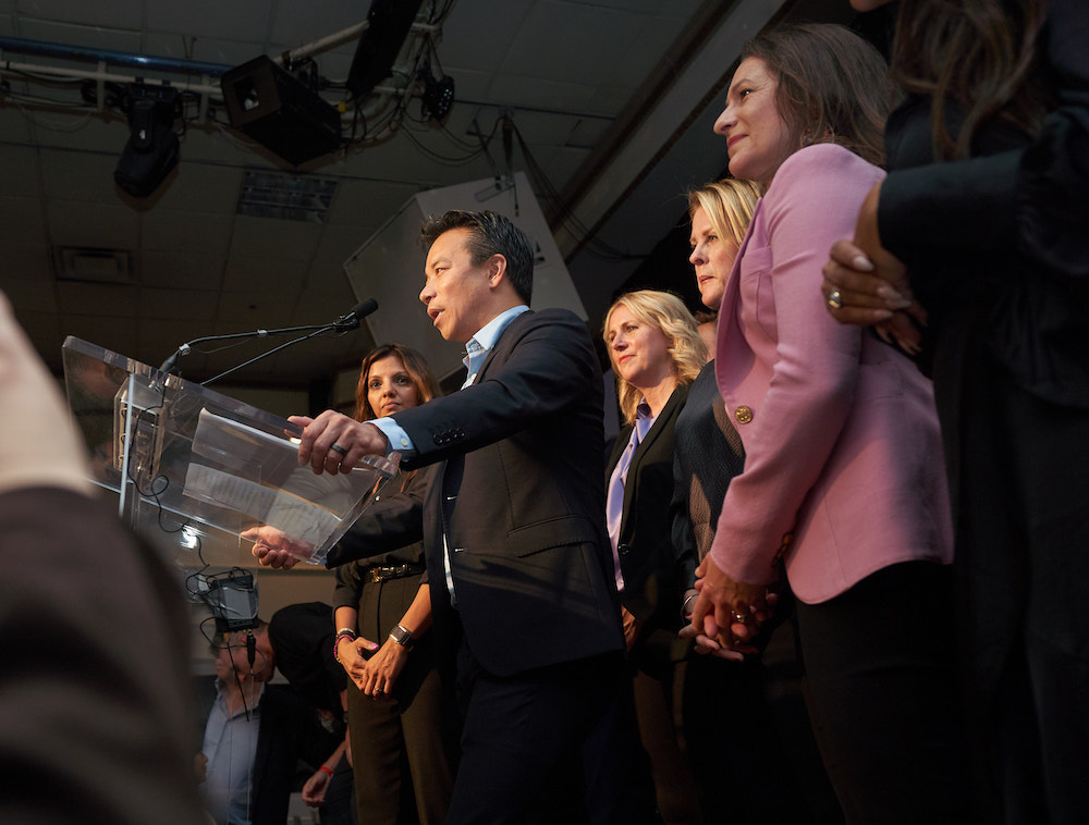 Ken Sim grips a clear glass podium while addressing the crowd at South Hall in Vancouver Oct. 15, 2022. He is wearing a light blue shirt under a black suit. Standing behind him are three newly-elected city councillors part of his ABC Vancouver party. From Sim’s right are Sarah Kirby-Yung (in a black blazer with a light blue shirt), Rebecca Bligh and Lisa Dominato (in a pink blazer).