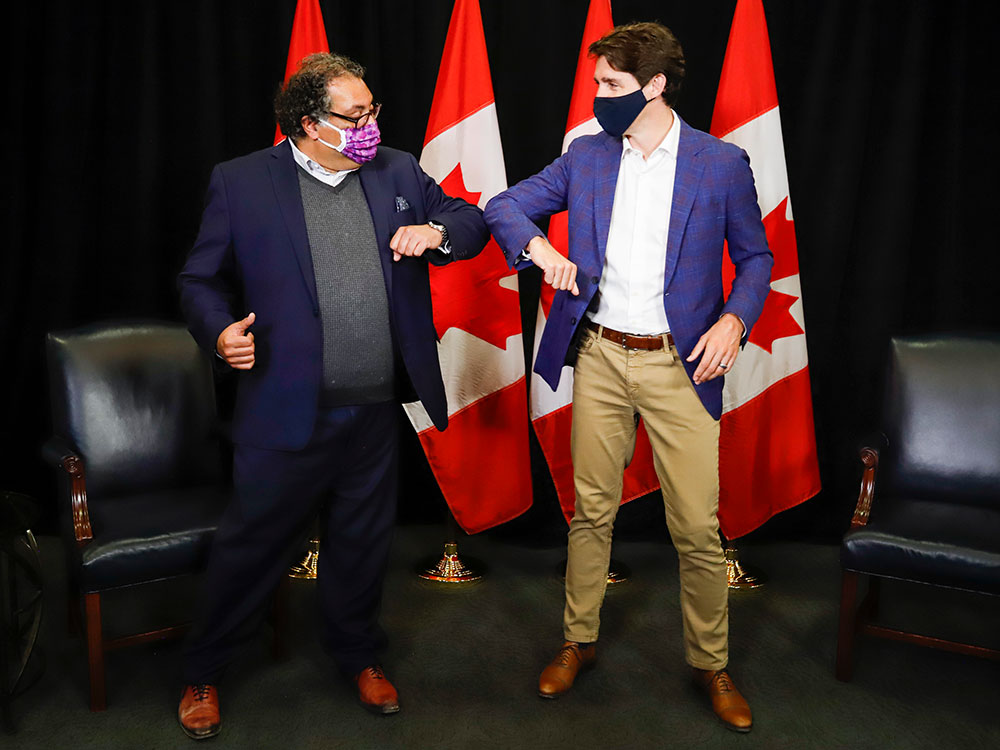 Trudeau and Nenshi, both wearing masks, bump elbows on a stage that features two chairs and four Canadian flags.