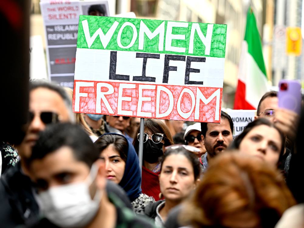 Amidst a crowd of protesters, a woman holds a sign that his hand coloured like the Iranian flag, with horizontal stripes of green, white and red, and over them the words Women, Life, Freedom.