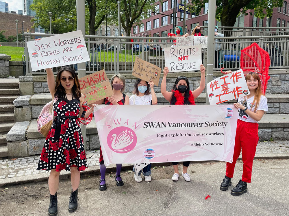 Five people stand at Victory Park in Vancouver. One person holds a red umbrella. Everyone holds signs reading things such as, “Sex Workers Rights Are Human Rights” and “Rights not Rescue.” Two signs feature Chinese characters. 