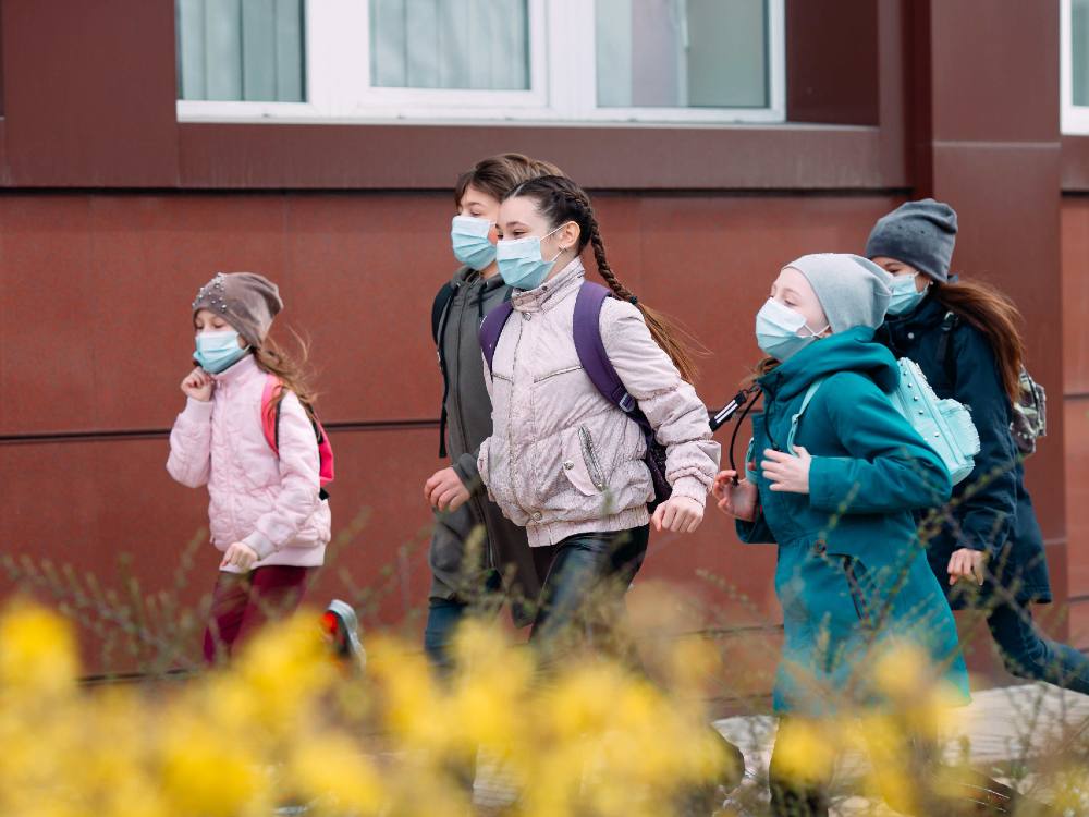 A group of children wearing masks, coats and toques run together in front of a building.