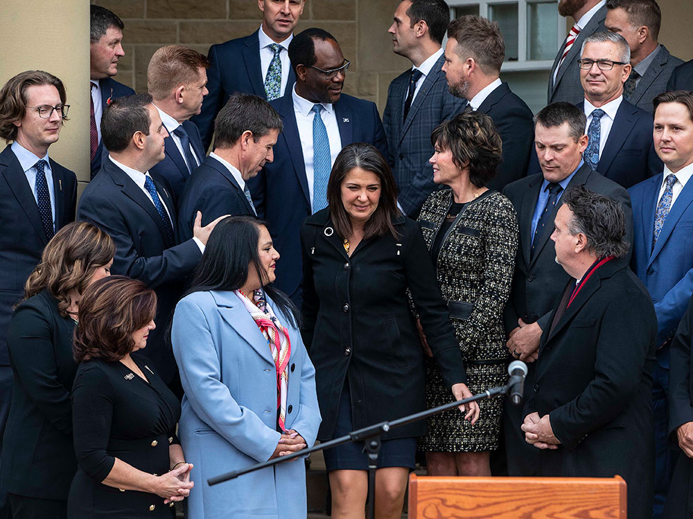 A crowd of people fills the frame. They are wearing dark suits — navy and black — and standing on a flight of outdoor stairs. Alberta premier Danielle Smith makes her way towards the podium at the bottom of the frame. She has dark shoulder-length hair and is wearing a black jacket and navy skirt. The people around her, her new cabinet members, are turned towards her and making space for her to descend the stairs.