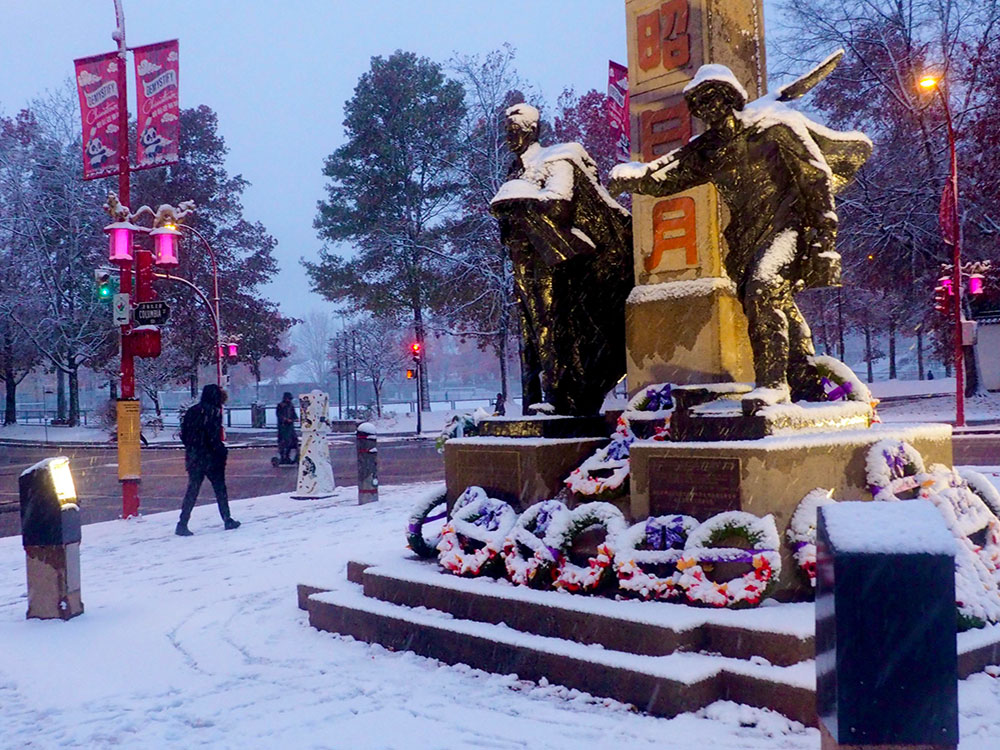 An early evening photo of a war memorial with a dusting of snow. Two sculptures stand on the podium, and snow-covered wreaths are arrayed in front of it. Pedestrians walk through the snow.