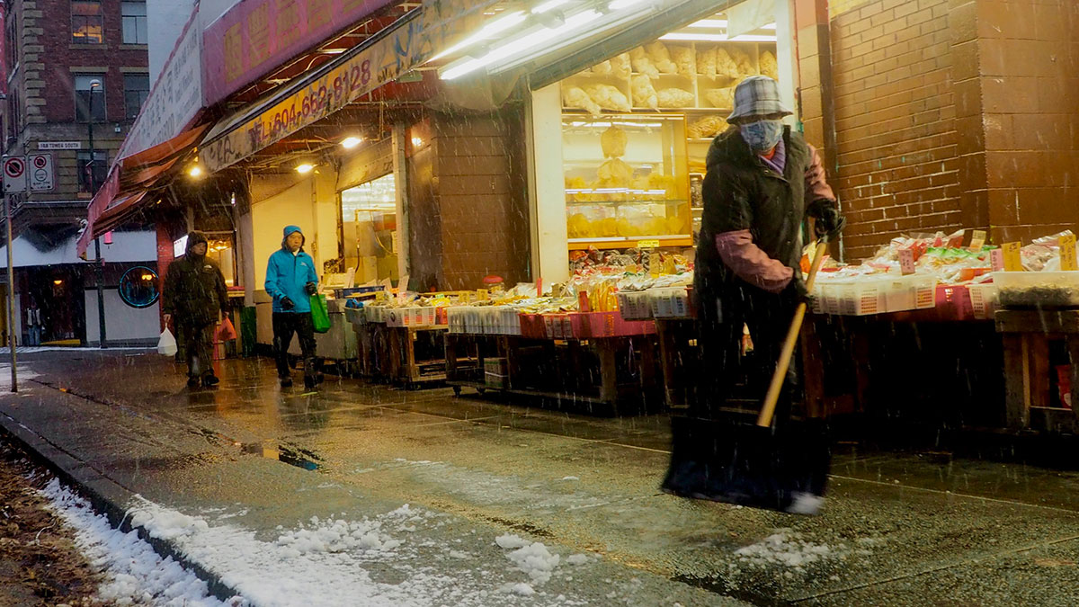 A person wearing a bulky coat and hat shovels a small amount of snow in front of a shop with a large open door and lighted interior. Two people are on the sidewalk.