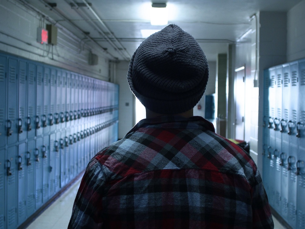 A teenager in a grey tuque and plaid shirt walks down a dimly lit hallway in a high school. There are rows of blue lockers on either side of them. Their back is turned to the camera. 
