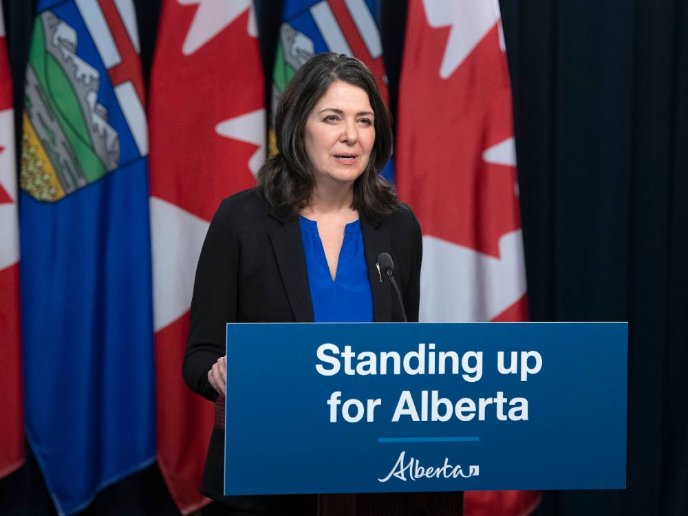 A woman with shoulder-length dark hair, wearing a medium blue shirt and dark blue jacket, stands at a podium with a blue sign saying “Standing up for Alberta”. Behind her is a row of Alberta and Canadian flags.