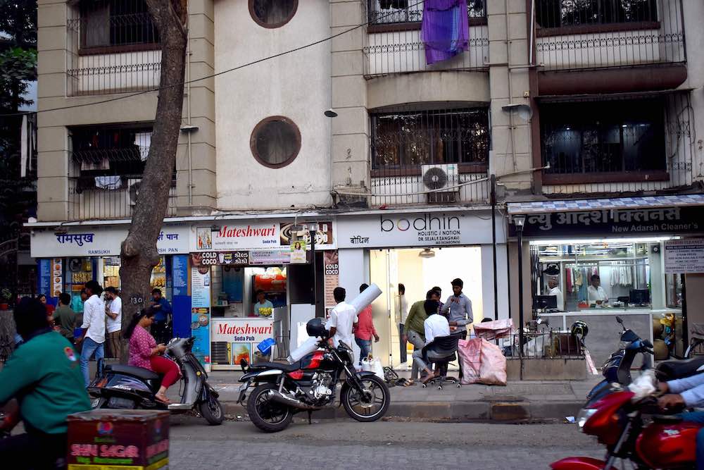 An evening streetscape featuring people on scooters in front of a line of storefronts connected to tall beige concrete apartment buildings.