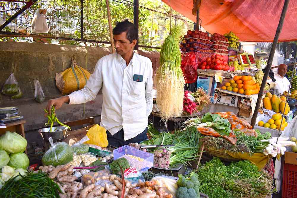 A food vendor in Mumbai is wearing a white button-down shirt. He is placing a handful of green chilis on a small stainless-steel scale. He is standing under a red awning and surrounded by fresh produce in an outdoor food stall.