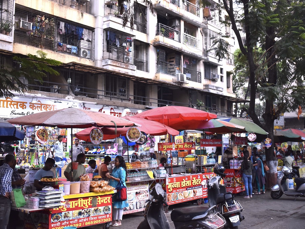A street scene in Mumbai depicts bustling food vendors under bright red umbrellas. The stalls are full of people and there are scooters in the foreground. The market stalls are on the street in front of a large white apartment building. Laundry is hanging on the square balconies.