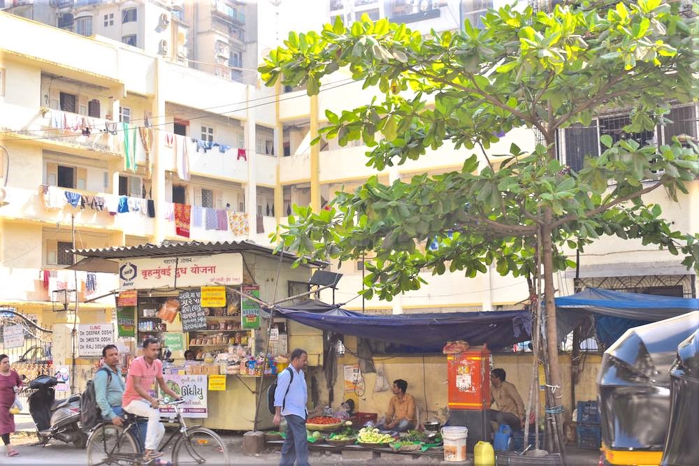 A streetscape of street vendors and cyclists against a light yellow high-rise apartment building in the sun, with clothing hanging from the balcony walls.