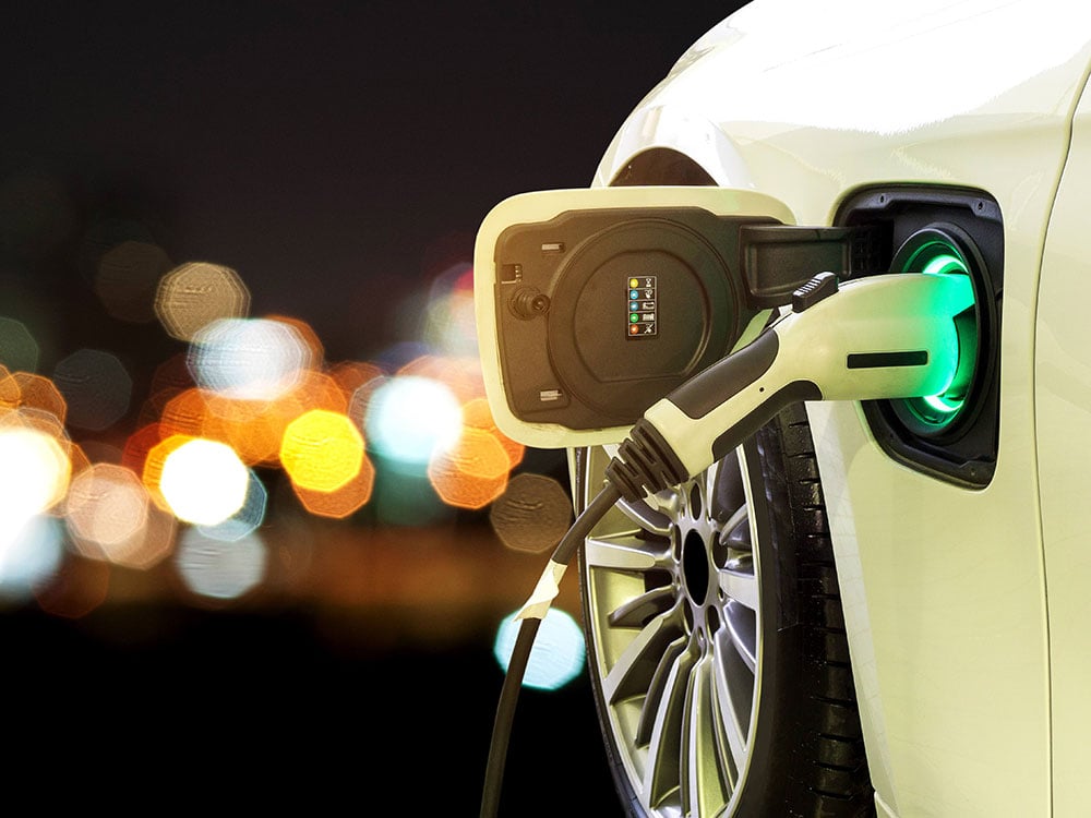 A nighttime photo of an electric vehicle charger installed in a white car to the right of the frame. There is a wash of blurry, colourful lights in the background.