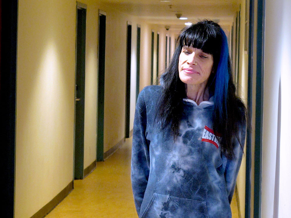 Julie Chapman is standing in a hallway. She has long black hair with a blue streak. She is looking down to the side. She is wearing a blue and white hoodie that says “East Van” on the side. 