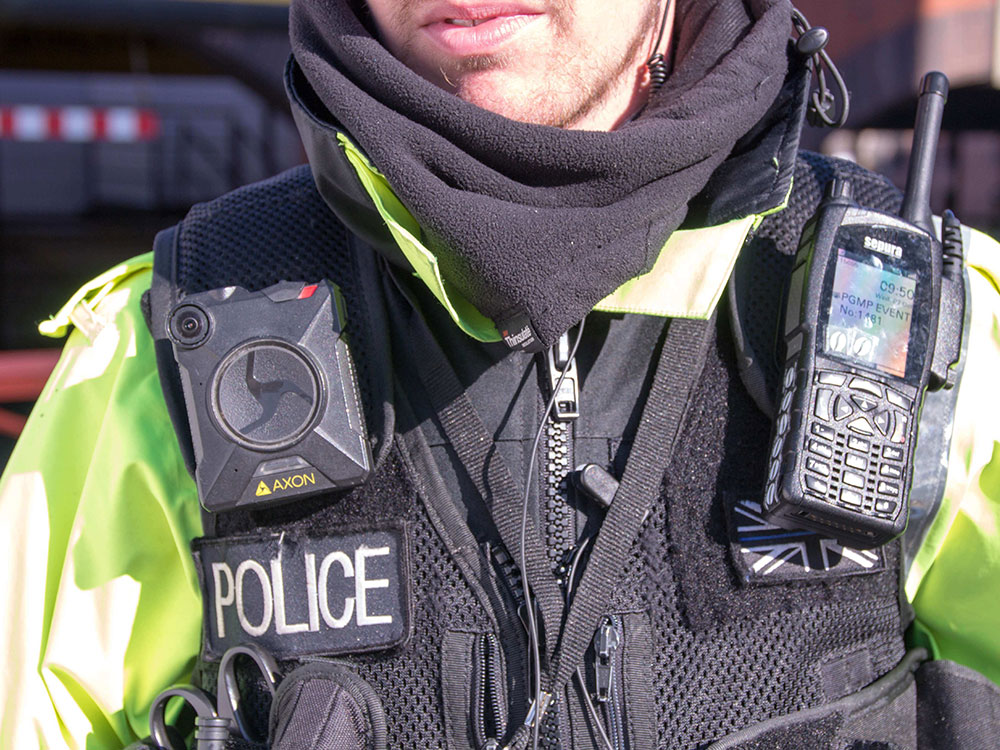 A Manchester police officer wears a fluorescent green shirt under a black vest equipped with a front-facing body camera on the left and a radio on the right. Their neck and mouth is visible, but the rest of their face is not.
