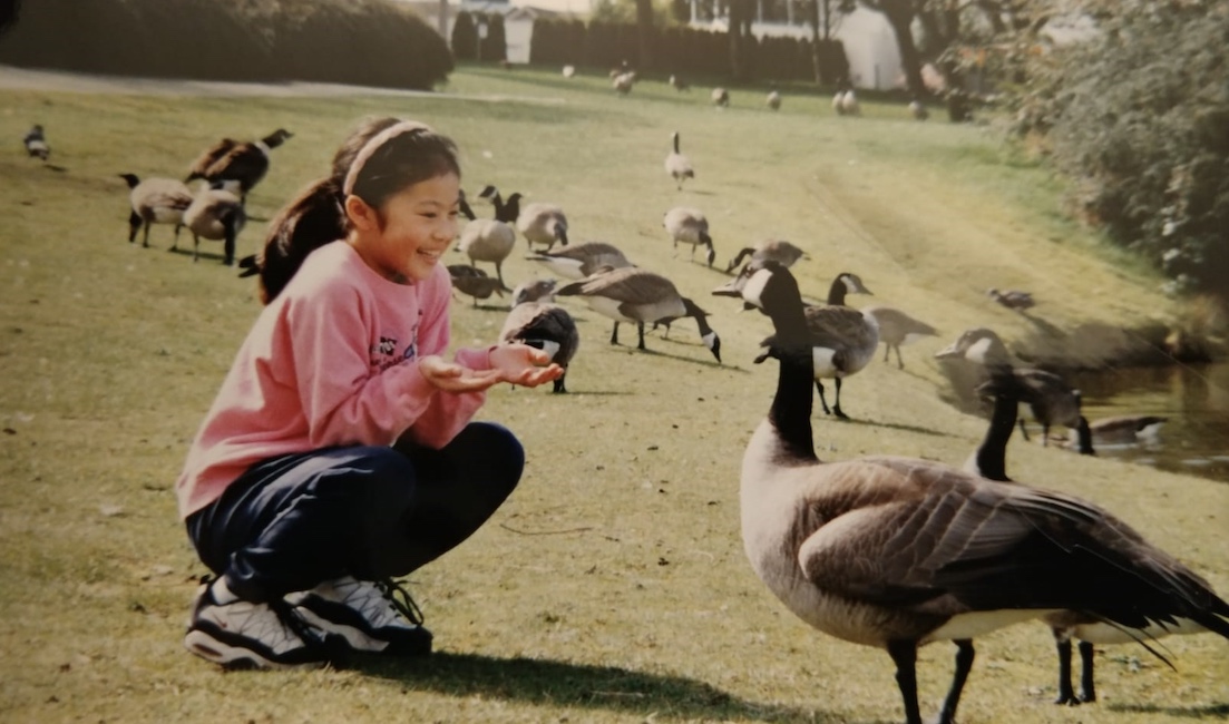 A photograph of Nicole Chan as a child. She is wearing a pink sweatshirt and crouching to the left of th frame near a Canada Goose in a sunny green field.