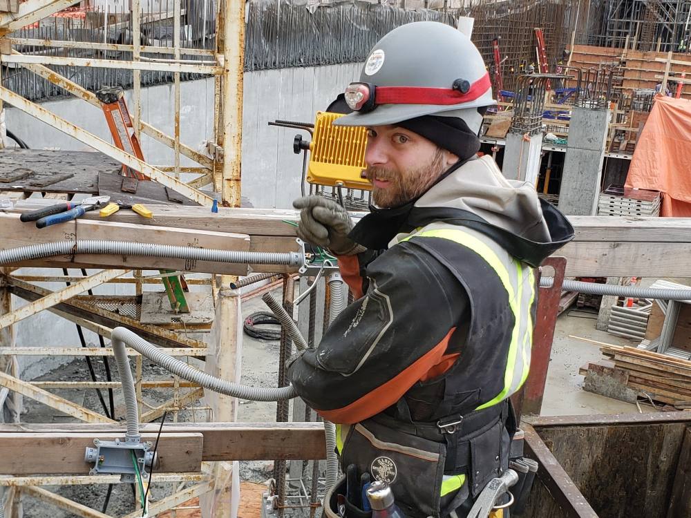 A young man with a beard and light grey construction helmet turns towards the camera behind him. He is smiling slightly and pointing to his left. He is wearing a black jacket under a black safety vest with fluorescent yellow reflectors and a tool belt. He is standing on a riser in a construction site. In front of him is a network of scaffolding and an underground concrete wall.