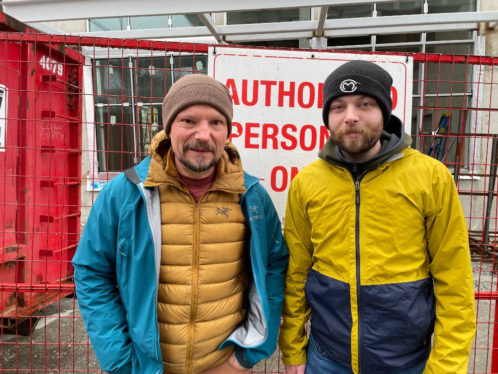 Two men stand in front of a construction site. They are standing in front of a red fence with a sign that reads “Authorized personnel only.” Nick Smith, left, is wearing a brown tuque and a yellow puffer jacket under a light blue rain shell. His son Emerson, right, is wearing a black tuque and a yellow and black rain jacket. Both are looking at the camera with neutral expressions.
