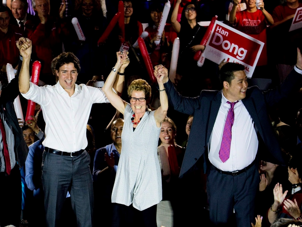 Prime Minister Justin Trudeau, in a white dress shirt with rolled-up sleeves, Kathleen Wynne, with glasses, short hair and a grey shirt, and Han Dong, wearing a blue suit and and purple tie, raise their hands together at a political rally.