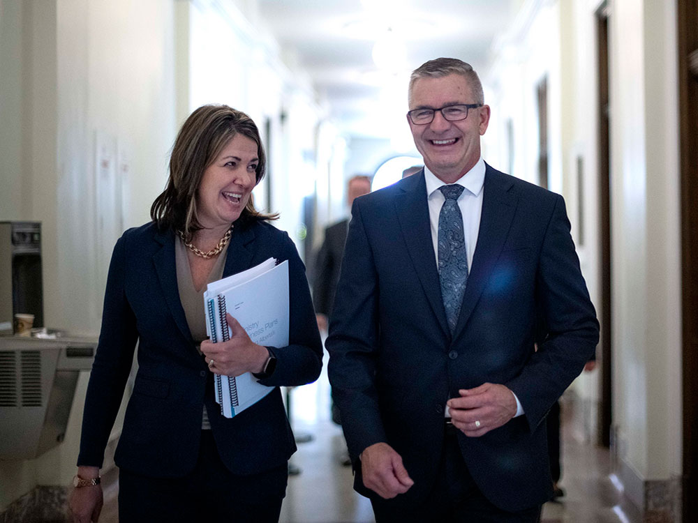 Alberta Premier Danielle Smith, wearing a black jacked and holding an armful of documents walks down a hall with Finance Minister Travis Toews who is in a dark suit, white shirt and tie. Both are smiling.