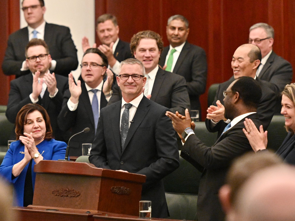 Finance Minister Travis Toews stands at a podium in the Legislative Assembly. He is surrounded by people, including Premier Danielle Smith, who are clapping for him. 
