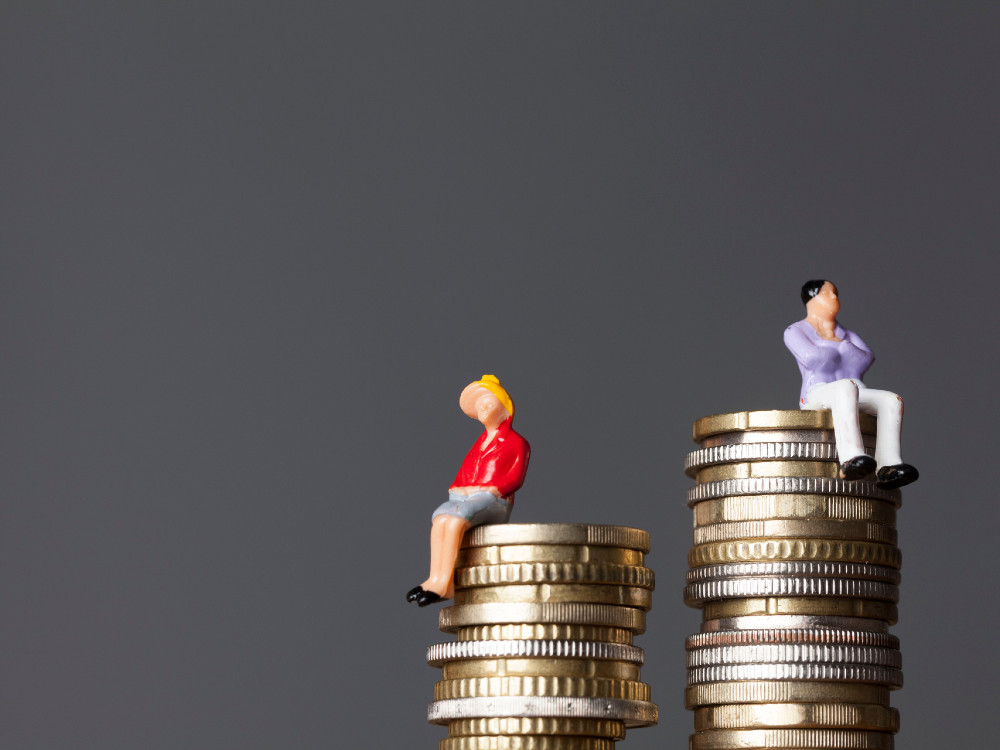 Two plastic miniature figurines sit atop a stack of gold and silver coins against a dark grey background. To the left, a woman with blonde hair, a red jacket and grey skirt sits on a shorter stack of coins. To the right, a man with black hair, a mauve jacket and white pants sits on a taller stack of coins. They are turned away from each other.  