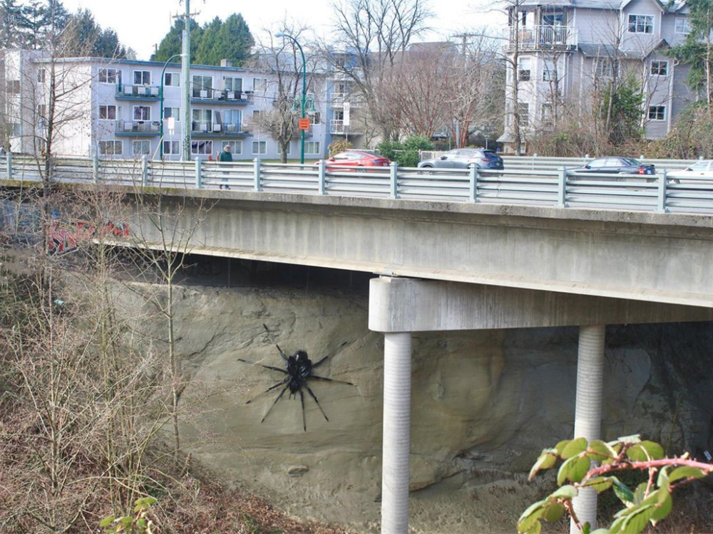 The photo shows a very large, shiny black spider sculpture on the concrete support for an overpass.