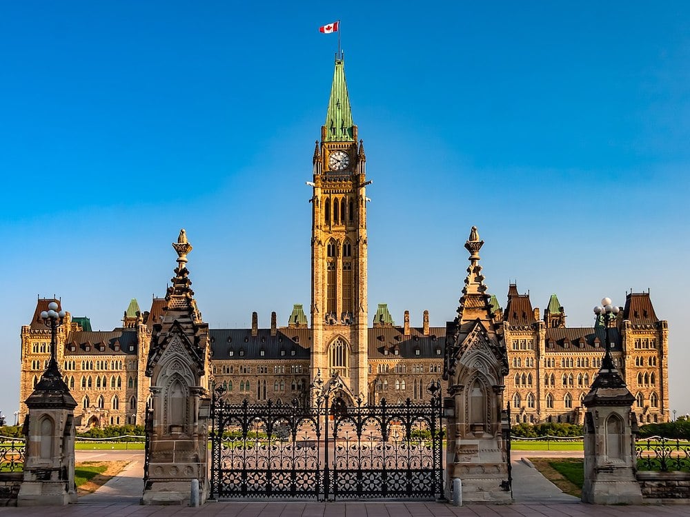 The gate to the Parliament buildings in Ottawa stand in shadow in front of the building, catching the sun amidst crisp blue sky.