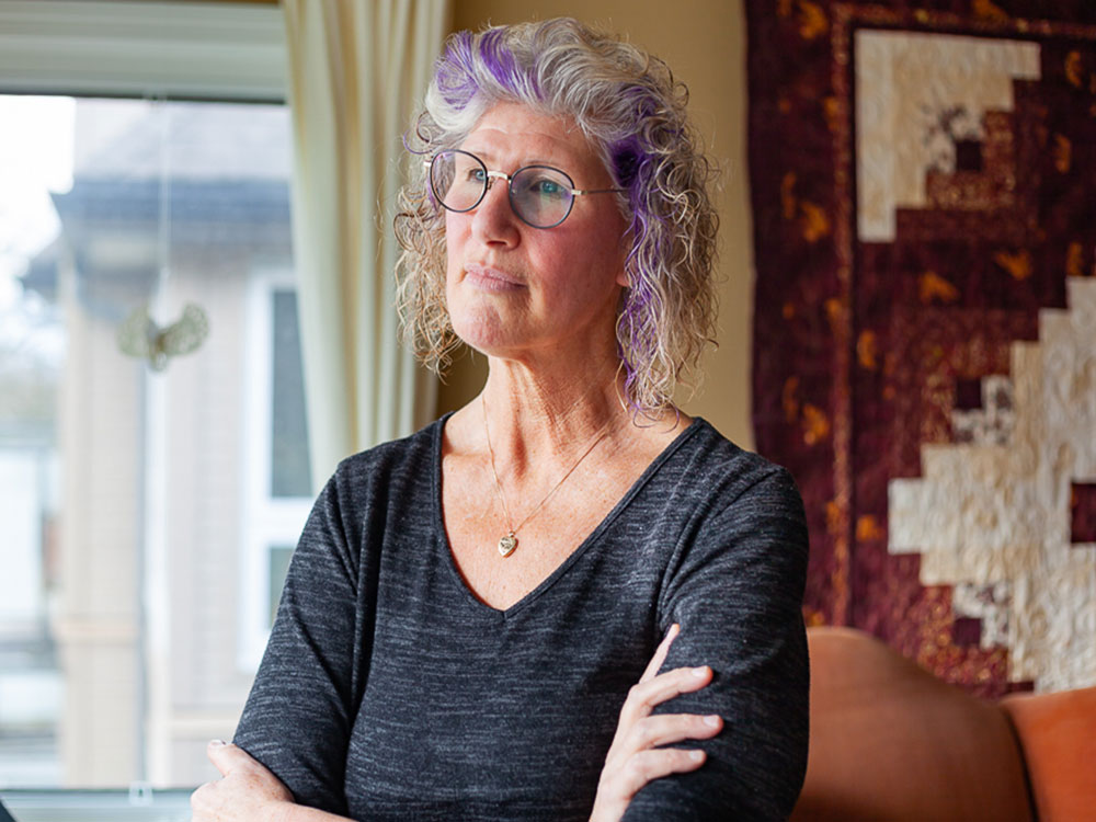 Tina Anderson has glasses and curly greying hair with a purple streak. She is wearing a grey cotton V-neck shirt and is looking away from the camera towards the left of the frame with her arms folded. She is standing in an indoor residential space and a piece of brown and white textile art hangs on the wall behind her.