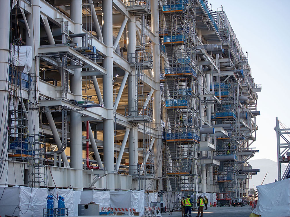 The photo shows the towering metal framework of the LNG Canada plant under construction.