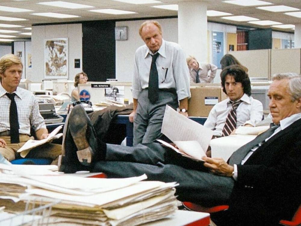 Movie actors, including Robert Redford, Dustin Hoffman and Jason Robards, play journalists in a Washington Post newsroom of the early 1970s, sitting or leaning on desks.
