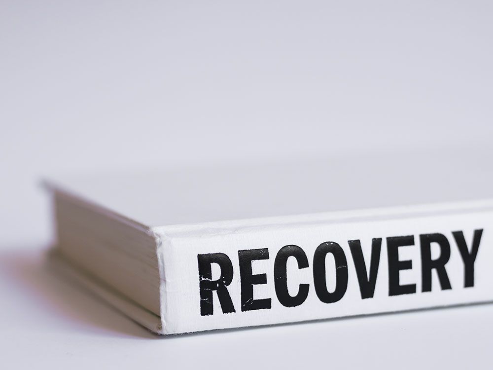 A white hardcover book reading “RECOVERY” in bold black letters along the spine sits amid a white background.