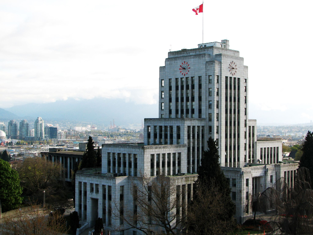 A view of the southwest side of Vancouver City Hall, a grey art deco building with a red neon clock near the top of its central tower. 