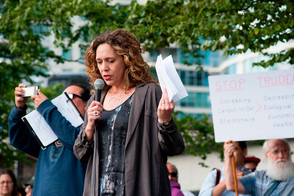 A woman with curly hair, holding sheets of paper and a microphone, addresses a crowd.