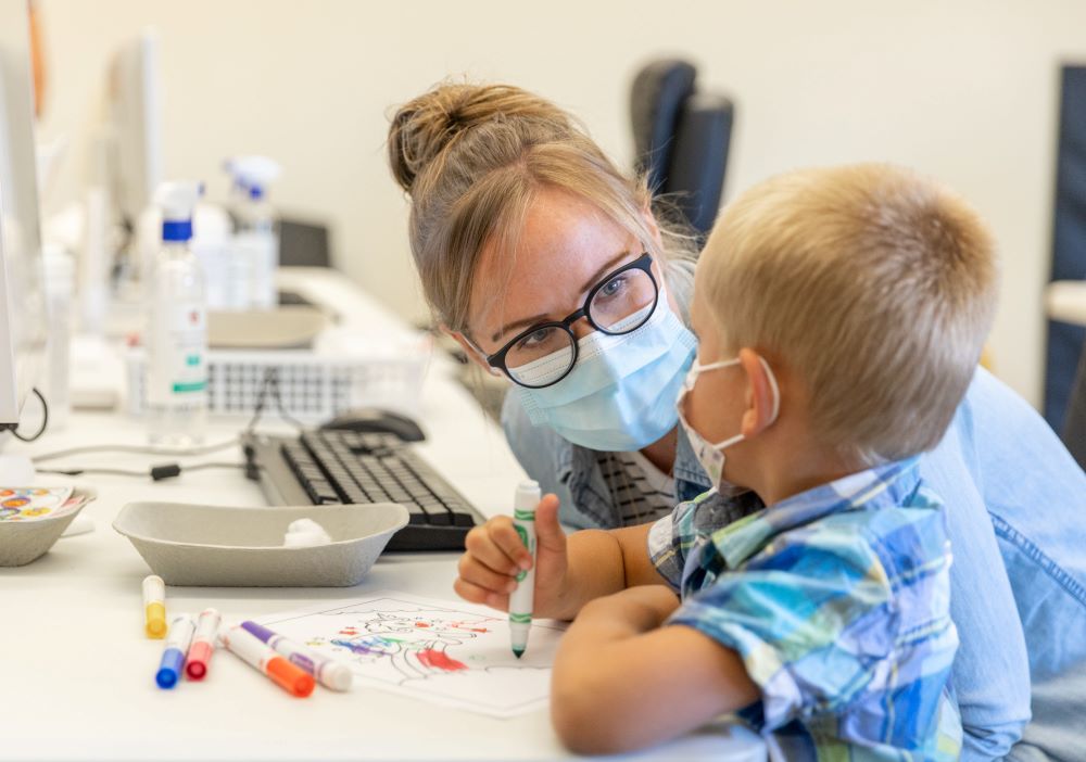 A woman with short blonde hair, glasses and light denim shirt is sitting at a table in a medical clinic and leaning towards a young child, looking at him warmly. The child has blonde hair and is wearing a plaid short-sleeved shirt. He is looking towards her and holding a green Crayola marker. They are both wearing medical masks.