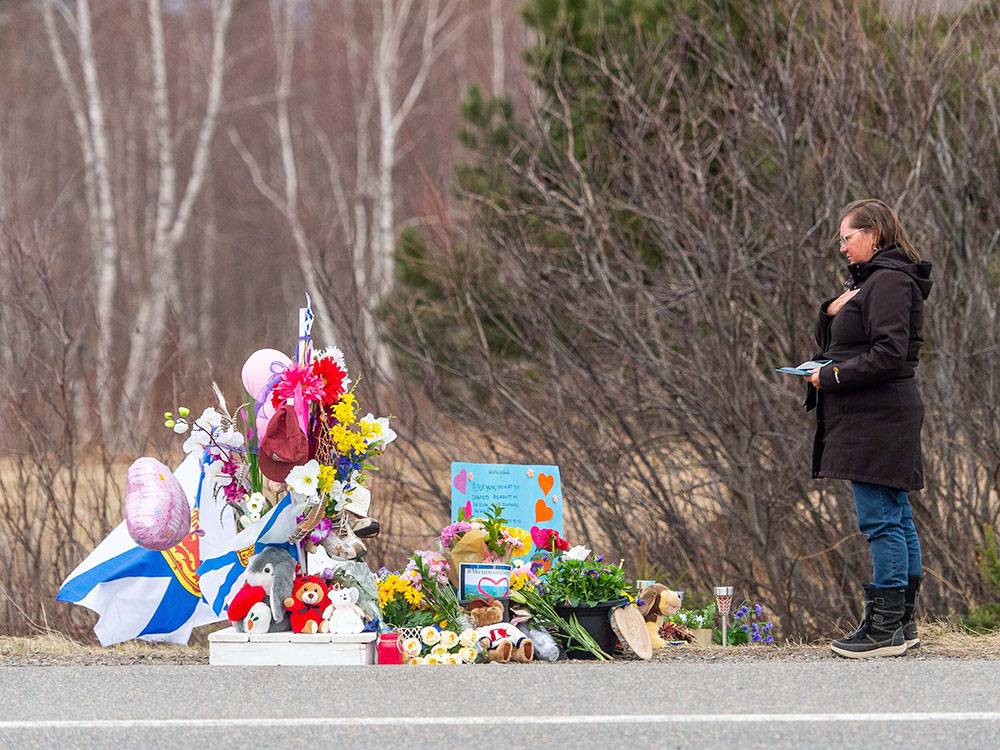 A person stands with hand on heart beside a memorial of flowers, a flag, toys and balloons.