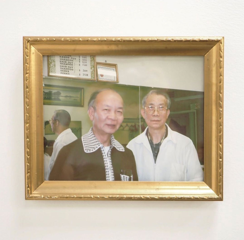 An old colour photo in a gold frame hangs on a white wall. It depicts Jack Gee, left, an elderly Chinese man in a brown cardigan, standing in a barber shop next to his friend Roy, right, in a white barber’s coat over a plaid shirt.