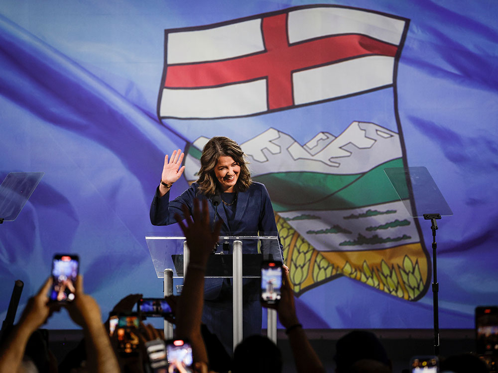 Danieille Smith, in a blue suit, waves to a crowd. She’s behind a podium and in front of a giant Alberta flag backdrop.