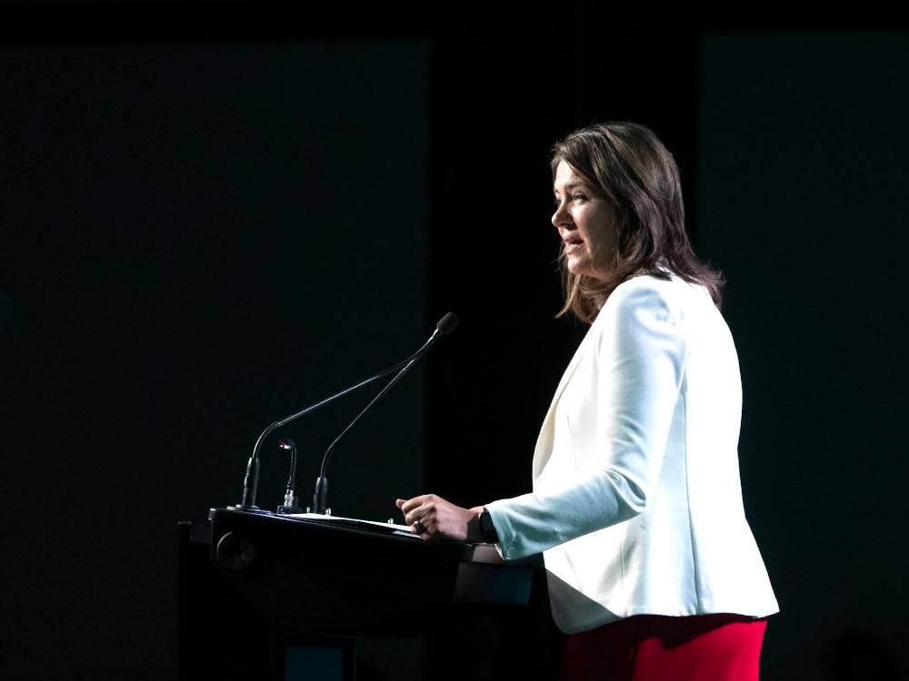 Danielle Smith, in a white jacket and red skirt, stands at a podium in a darkened room.
