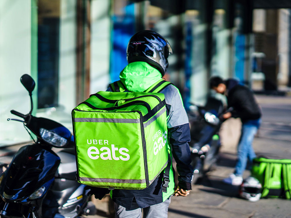 A person wearing a helmet walks on a sidewalk with a large green backpack labelled Uber Eats.